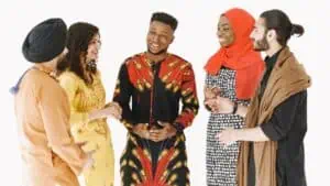 Cutural Intelligence. The image showcases a group of five diverse individuals standing together against a white background. They appear to be engaged in a lively conversation, smiling and displaying positive body language. From left to right: A person wearing a turban, dressed in an orange outfit with a blue tassel. A woman wearing a vibrant yellow dress adorned with intricate designs. A man in a striking red and black patterned outfit with motifs resembling traditional African prints. He is laughing and has a joyful expression. A woman wearing a red hijab and a black and white patterned dress. A man with long hair tied back, wearing a brown cloak or shawl. The group seems to represent a mix of cultures and backgrounds, emphasizing unity and diversity.