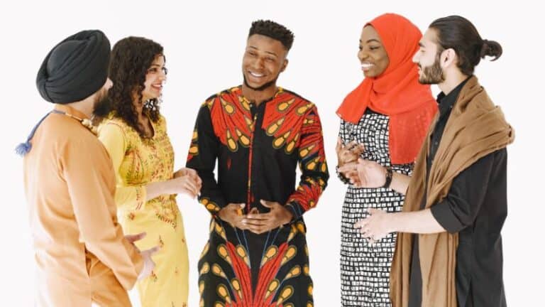Cutural Intelligence. The image showcases a group of five diverse individuals standing together against a white background. They appear to be engaged in a lively conversation, smiling and displaying positive body language. From left to right: A person wearing a turban, dressed in an orange outfit with a blue tassel. A woman wearing a vibrant yellow dress adorned with intricate designs. A man in a striking red and black patterned outfit with motifs resembling traditional African prints. He is laughing and has a joyful expression. A woman wearing a red hijab and a black and white patterned dress. A man with long hair tied back, wearing a brown cloak or shawl. The group seems to represent a mix of cultures and backgrounds, emphasizing unity and diversity.