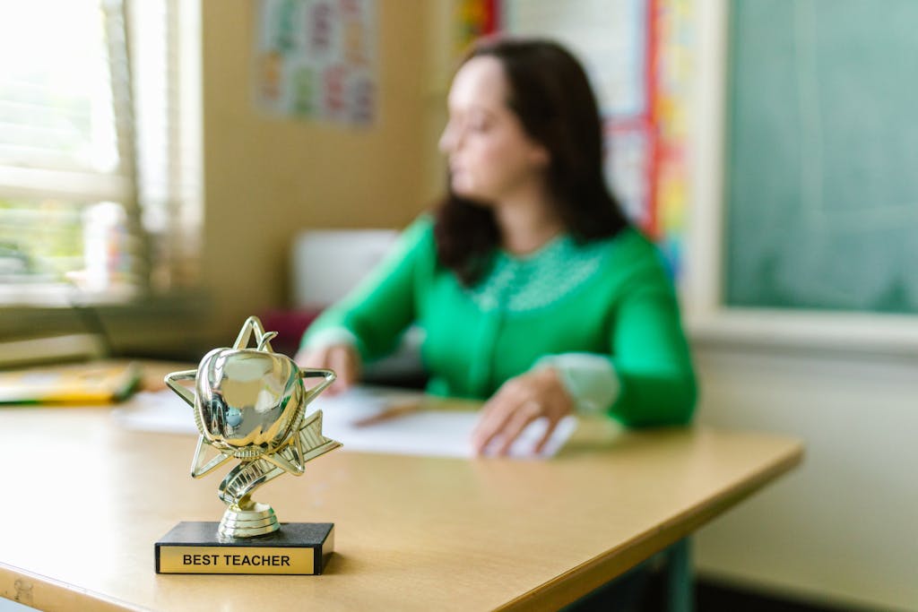 A blurred teacher in a classroom with a 'Best Teacher' trophy on the desk.