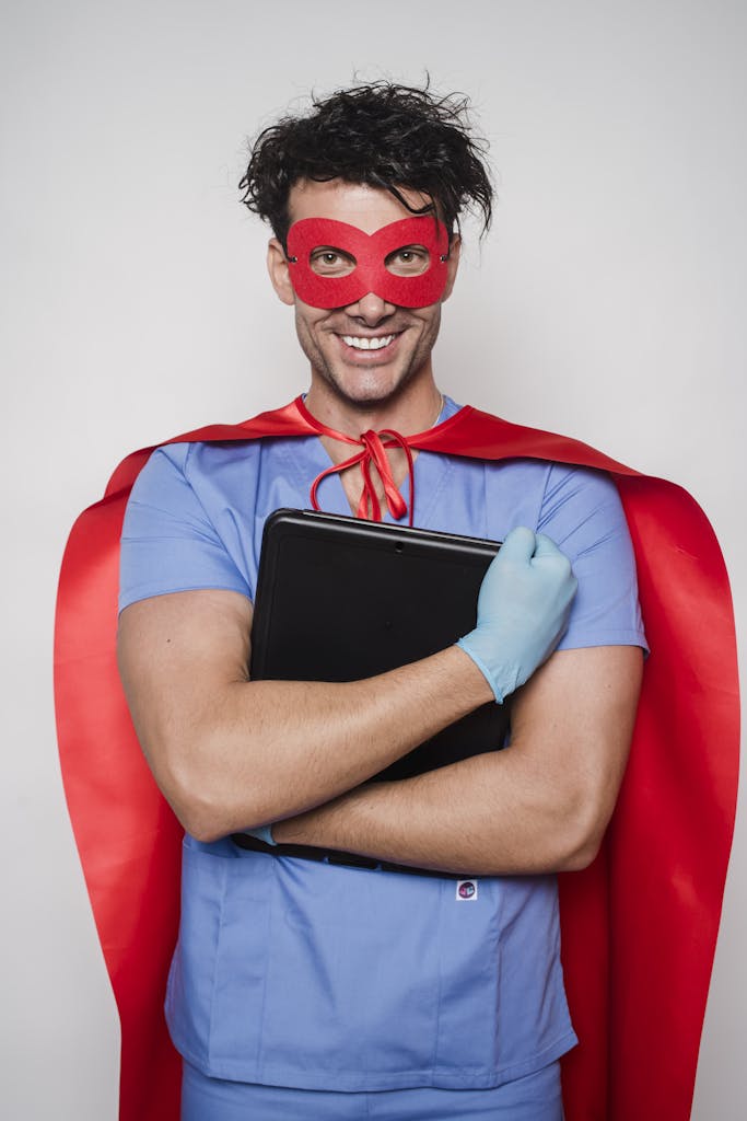 Cheerful man doctor in superhero costume with mask and cape standing with folder with documents and looking at camera against light wall