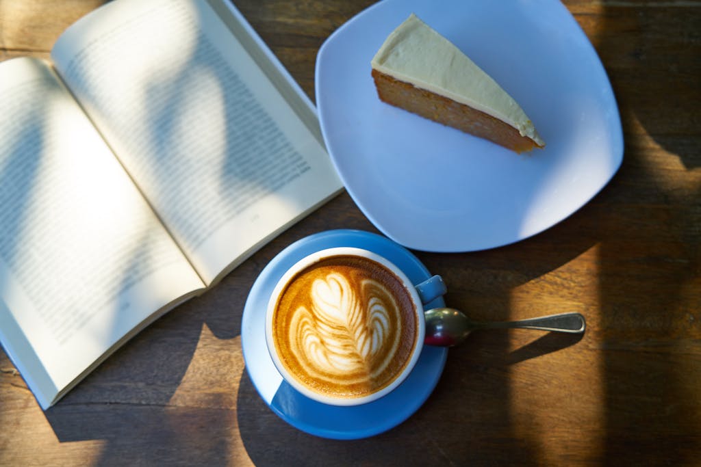 Photo by Pixabay A beautifully crafted latte with latte art, alongside a slice of cake and an open book on a wooden table.