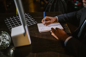 A businessman in a suit writing notes on a notebook at a desk, suggesting a professional office environment.