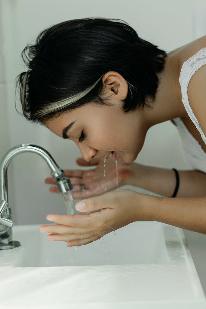 Photo by Vitória Santos A woman with short hair gently washes her face at a bathroom sink, embracing a clean and refreshing start to the day.