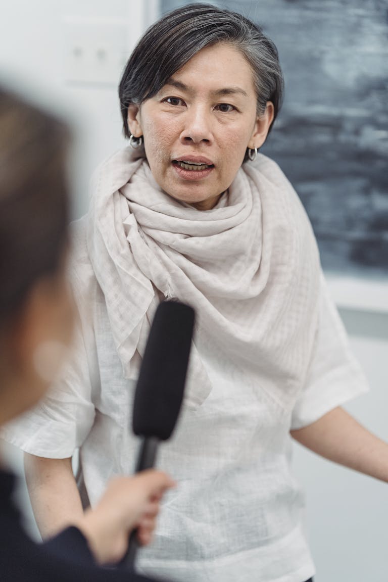 Mature woman speaking into a microphone, captured in a close indoor interview setting.