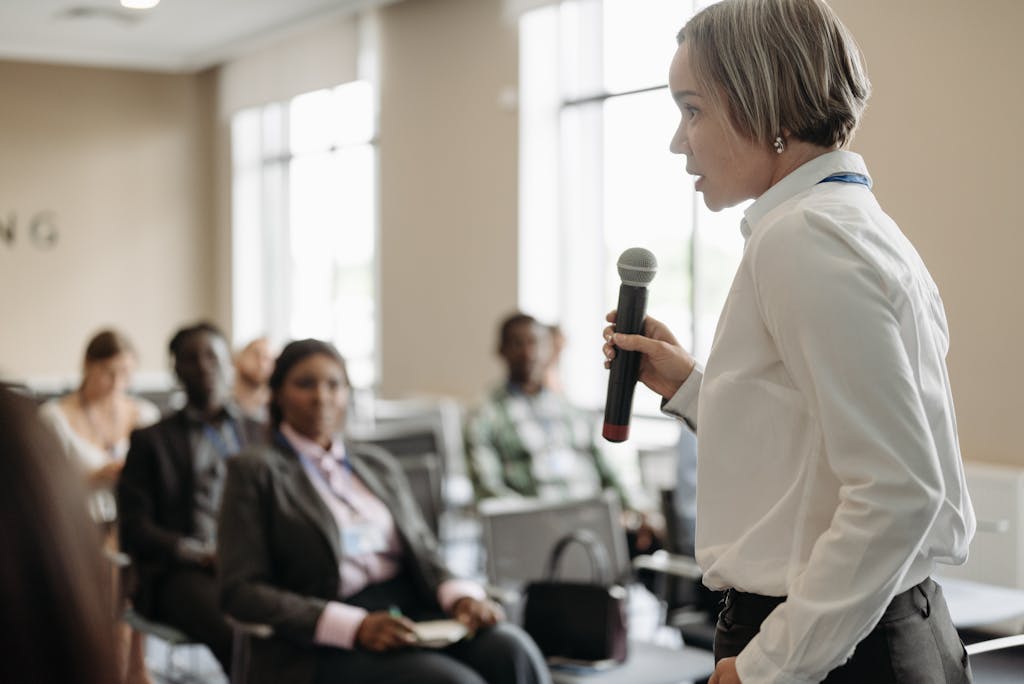 Photo by Pavel Danilyuk Side view of a businesswoman holding a microphone while addressing an audience at a conference.