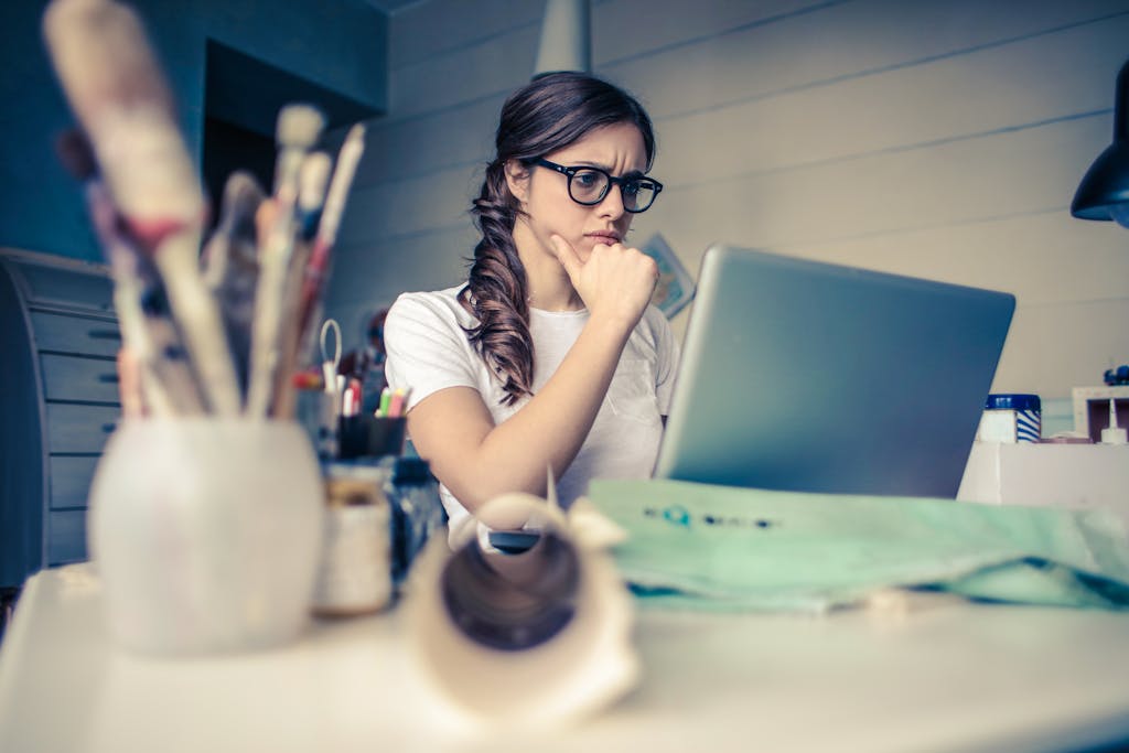 Photo by Andrea Piacquadio Young woman with glasses deeply focused on a laptop surrounded by art supplies in a home office.