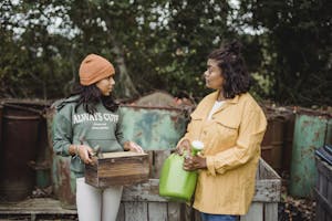 Ethnic woman with watering can speaking with teen near metal containers while looking at each other on farm