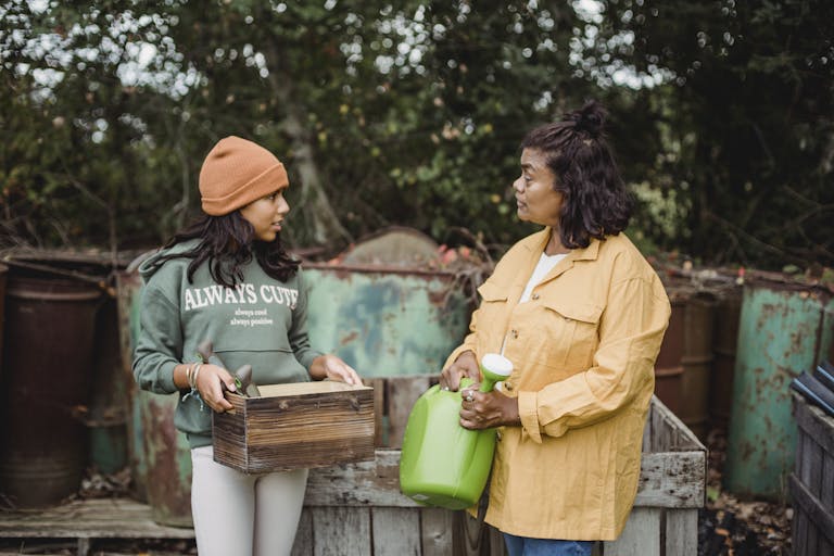 Ethnic woman with watering can speaking with teen near metal containers while looking at each other on farm