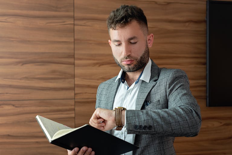 Young businessman in gray suit checking wristwatch while holding a notebook indoors.