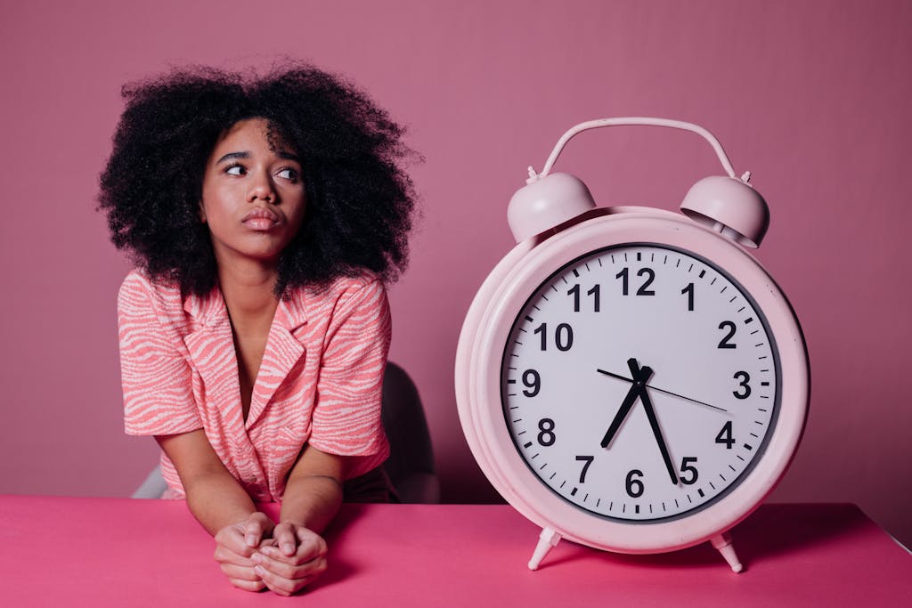 Pensive woman with afro hair leaning on a table next to a large pink alarm clock.