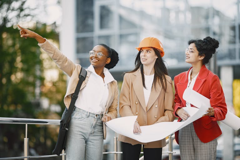 Three female professionals discussing construction plans outdoors with a city backdrop.